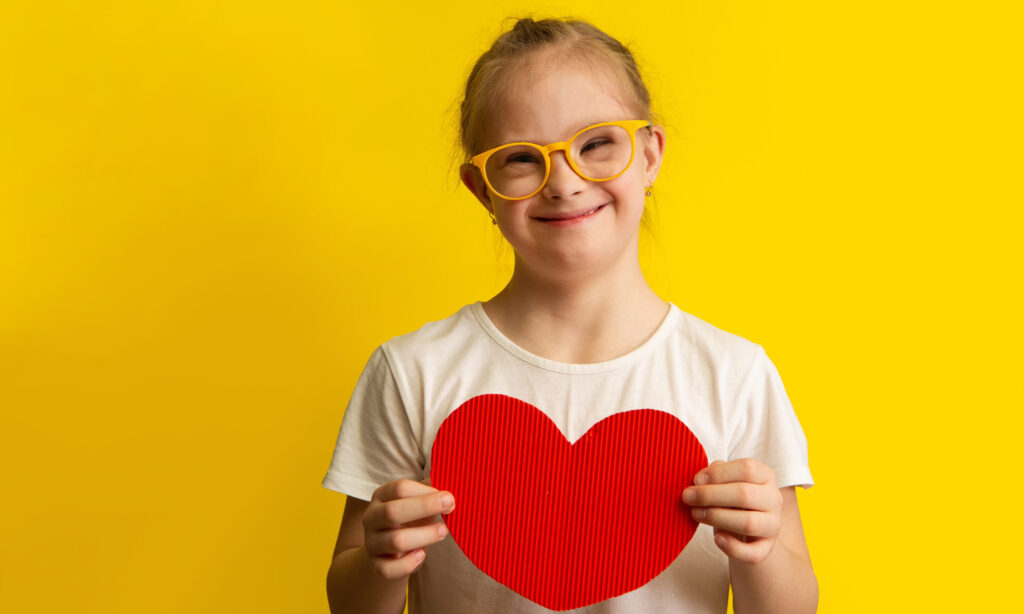 Girl with Down Syndrome holding a big red paper heart on a yellow background